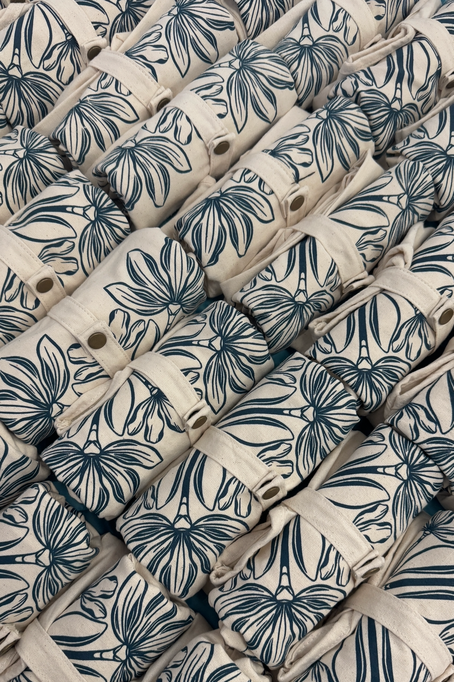 A close-up of multiple rolled totes stacked together, featuring a navy blue floral hawaiian quilt pattern on a warm off-white background.